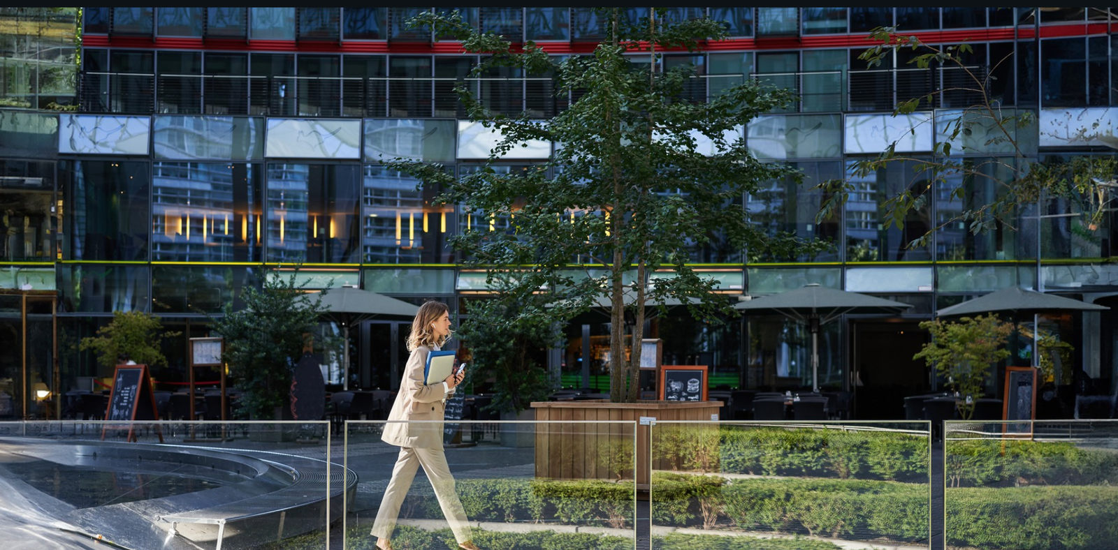 A professional woman walks past the outdoor area of a modern glass office building, representing Lets Connect’s hybrid-ready office spaces designed to support flexible work environments and set the brand apart for remote and on-site teams.